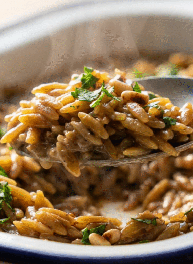 Spoon lifting creamy black garlic orzo from a baking dish, garnished with parsley and pine nuts
