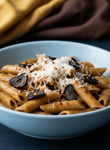 Black garlic penne pasta in a light blue ceramic bowl, topped with sliced black garlic, grated Parmesan, and chili flakes